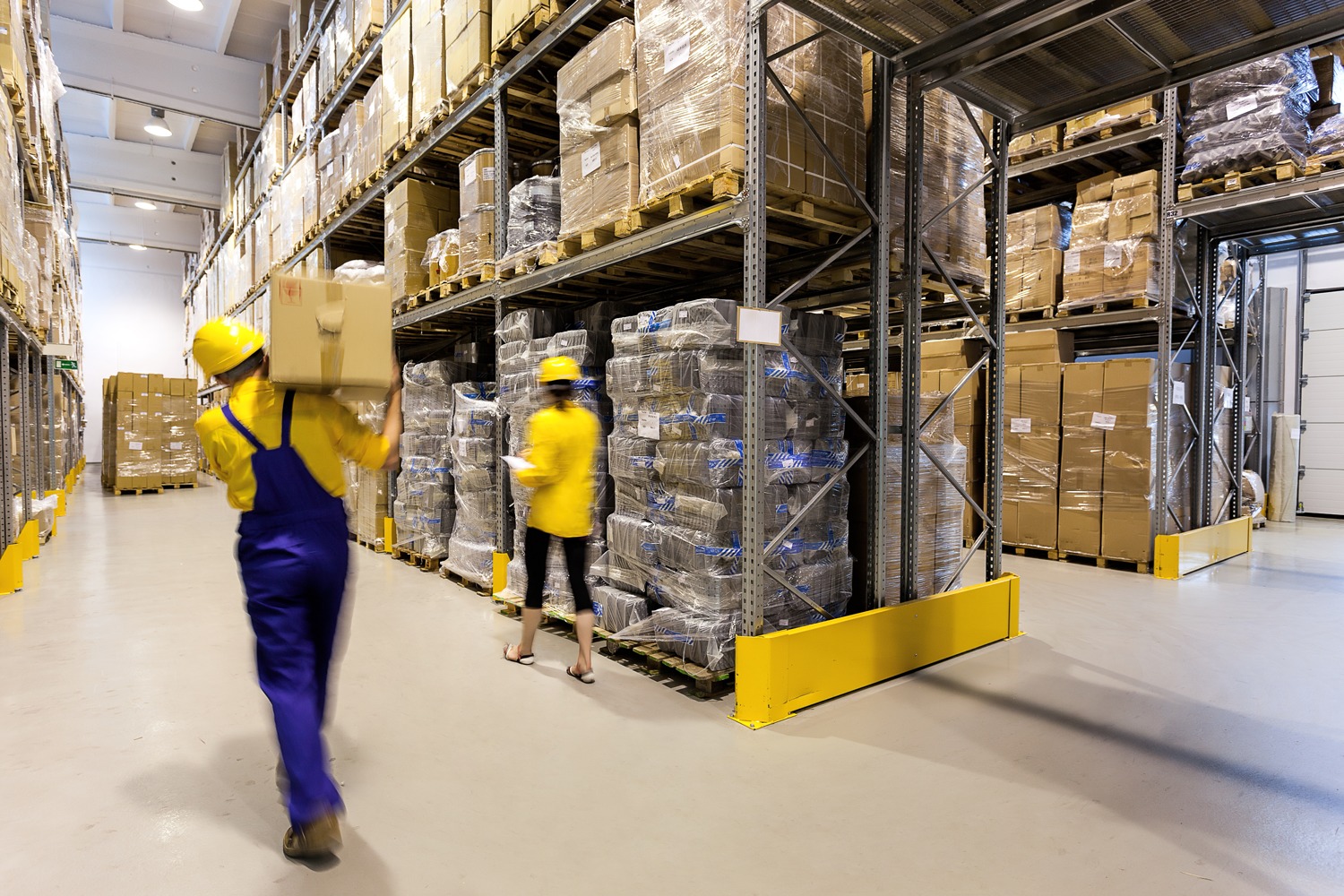 Two workers wearing yellow helmets and uniforms move boxes in a brightly lit warehouse filled with shelves stacked with pallets of goods and cardboard boxes.
