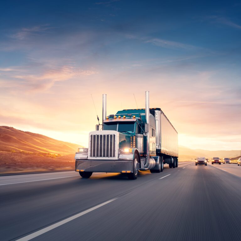 A large semi-truck drives down a multi-lane highway at sunset, surrounded by hills and other vehicles under a colorful sky.