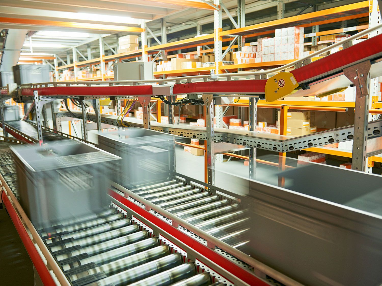 Plastic bins move quickly along a conveyor belt system in a warehouse, with shelves of boxes and packages in the background—showcasing an automated logistics center ideal for advanced automation analysis.