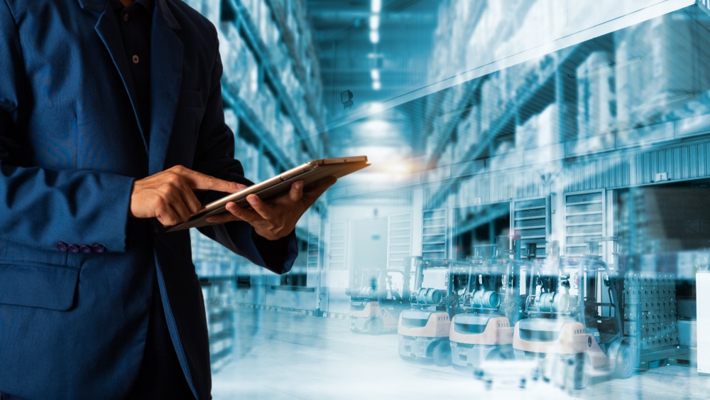 A person in a blue suit uses a digital tablet in a warehouse setting, with rows of shelves and forklifts in the background, representing logistics, inventory management, and supply chain planning.