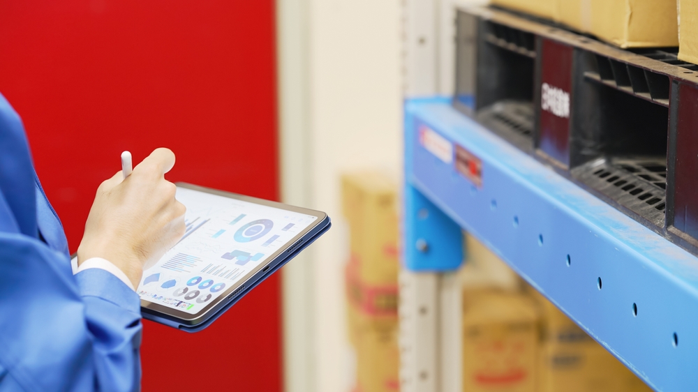 A person in a blue uniform holds a tablet displaying graphs and charts, using a WMS to conduct inventory or inspection in a warehouse filled with shelves and cardboard boxes.