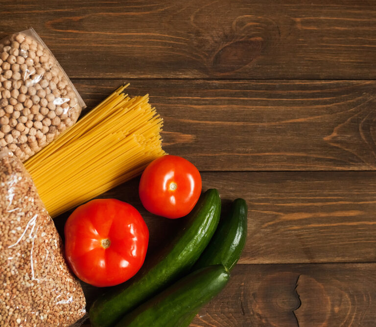 Packets of chickpeas, pasta, and grains, along with three tomatoes and three cucumbers, are arranged on a wooden table with empty space on the right.
