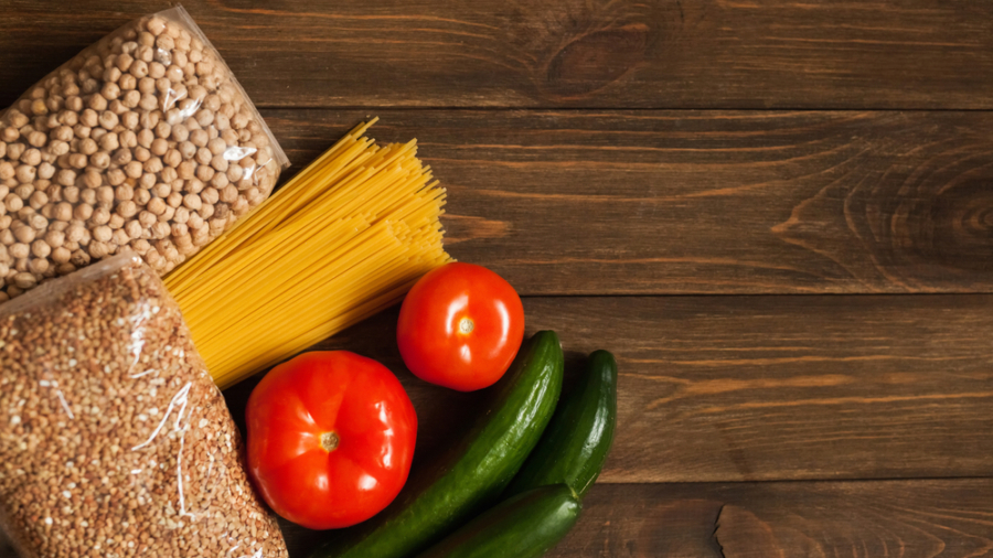 Packets of chickpeas, pasta, and grains, along with three tomatoes and three cucumbers, are arranged on a wooden table with empty space on the right.