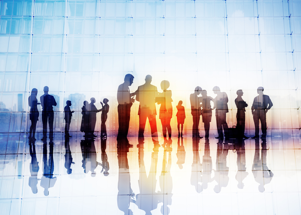 Silhouettes of business people discussing S&OP strategies in groups by large glass windows, their reflections on the shiny floor and a cityscape visible through the glass.