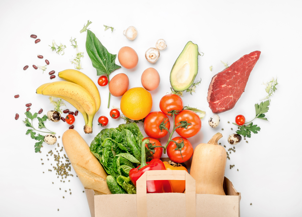 A paper bag overflows with fresh food—tomatoes, lettuce, bananas, bell pepper, baguette, eggs, and more—showcasing the importance of supply chain traceability on a white background.