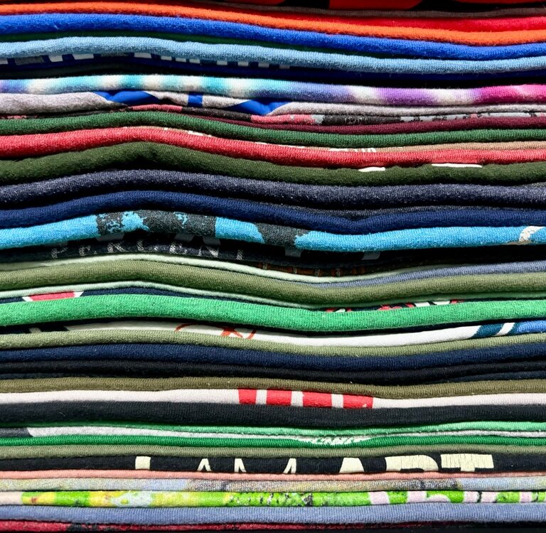 A close-up view of a neatly stacked pile of colorful folded t-shirts, arranged in horizontal layers showing various patterns and textures, highlights the variety available despite recent tariff changes.