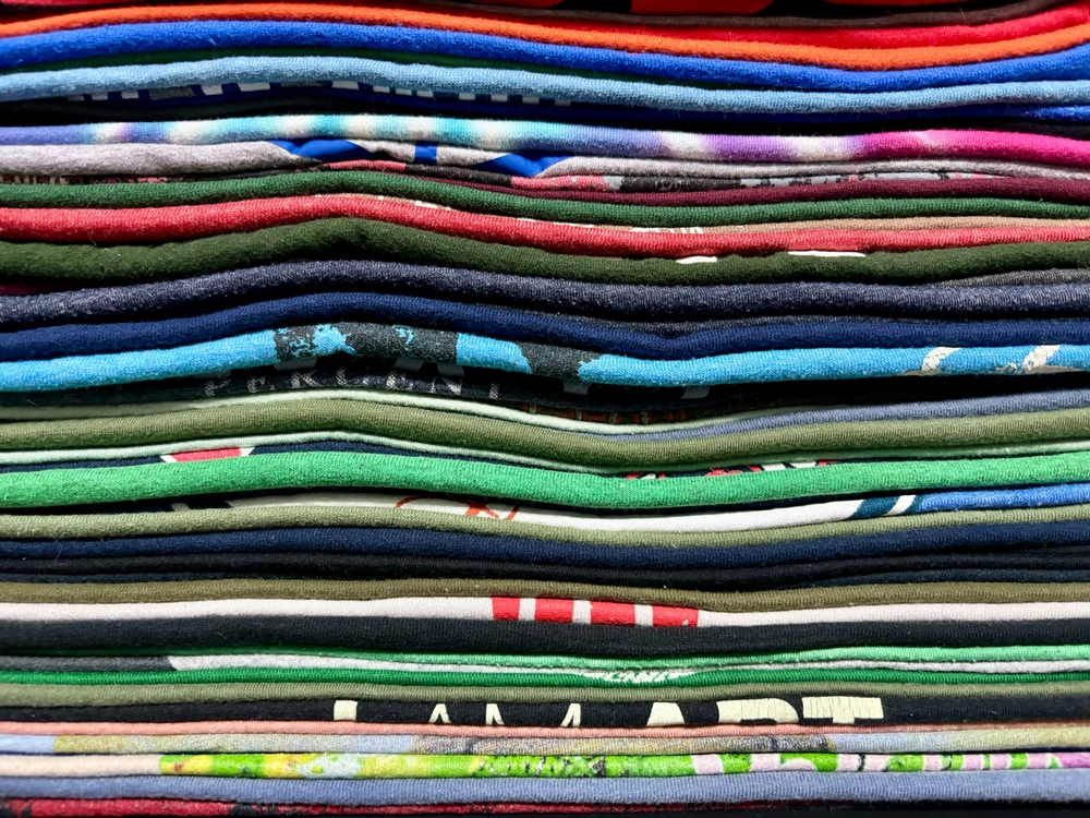 A close-up view of a neatly stacked pile of colorful folded t-shirts, arranged in horizontal layers showing various patterns and textures, highlights the variety available despite recent tariff changes.