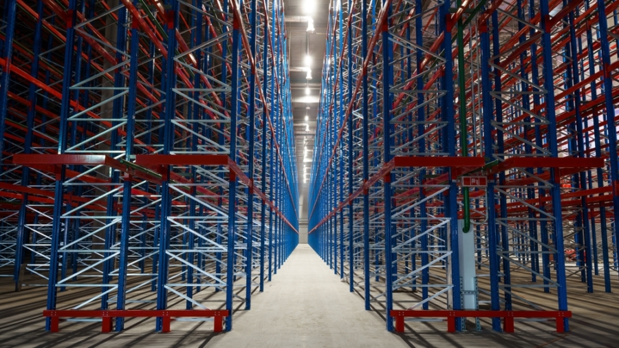 Rows of tall, empty metal storage racks in a brightly lit warehouse, forming a symmetrical, industrial corridor—standing ready for inventory as tariff changes impact the flow of goods.