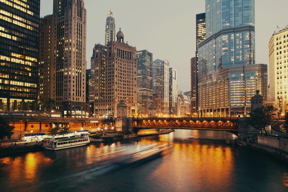 Cityscape at dusk showing tall skyscrapers reflected in a river, with a lit-up bridge spanning the water. Boats move along the river, leaving blurred trails, and lights glow from buildings and along the waterfront.