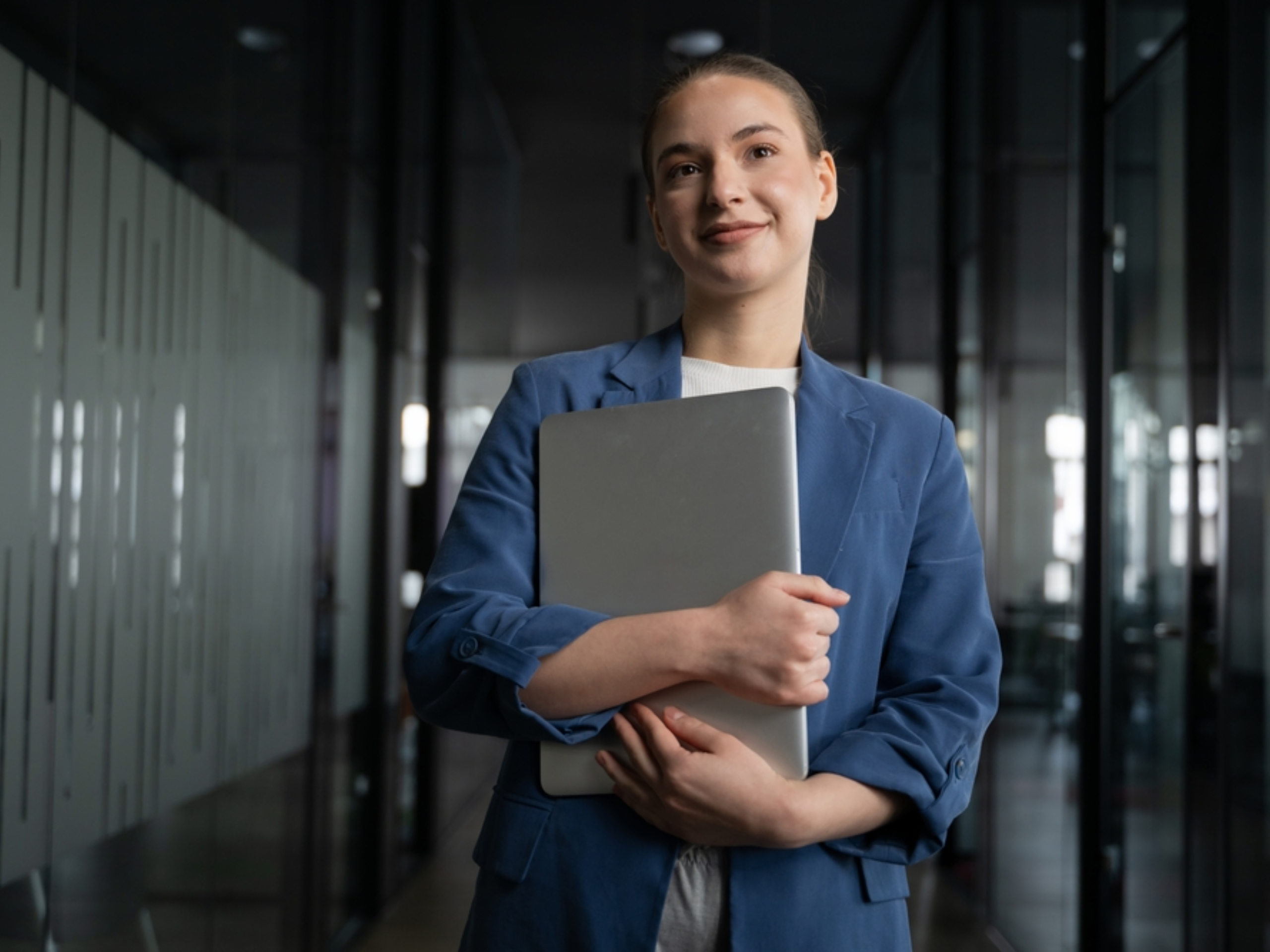 A young woman wearing a blue blazer stands in a modern office hallway, holding a closed laptop to her chest and smiling slightly, with glass walls and reflections in the background.