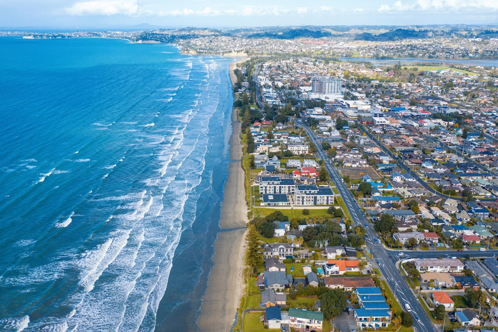 Aerial view of a coastal town with houses and roads lining a sandy beach, waves rolling onto the shore, and the ocean stretching to the horizon under a partly cloudy sky.