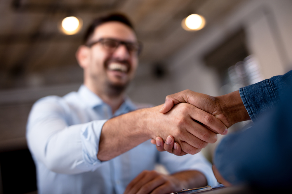 Close-up of two people shaking hands, with one person in focus and the other blurred in the background, smiling and wearing glasses, suggesting a positive and friendly interaction in a professional setting.