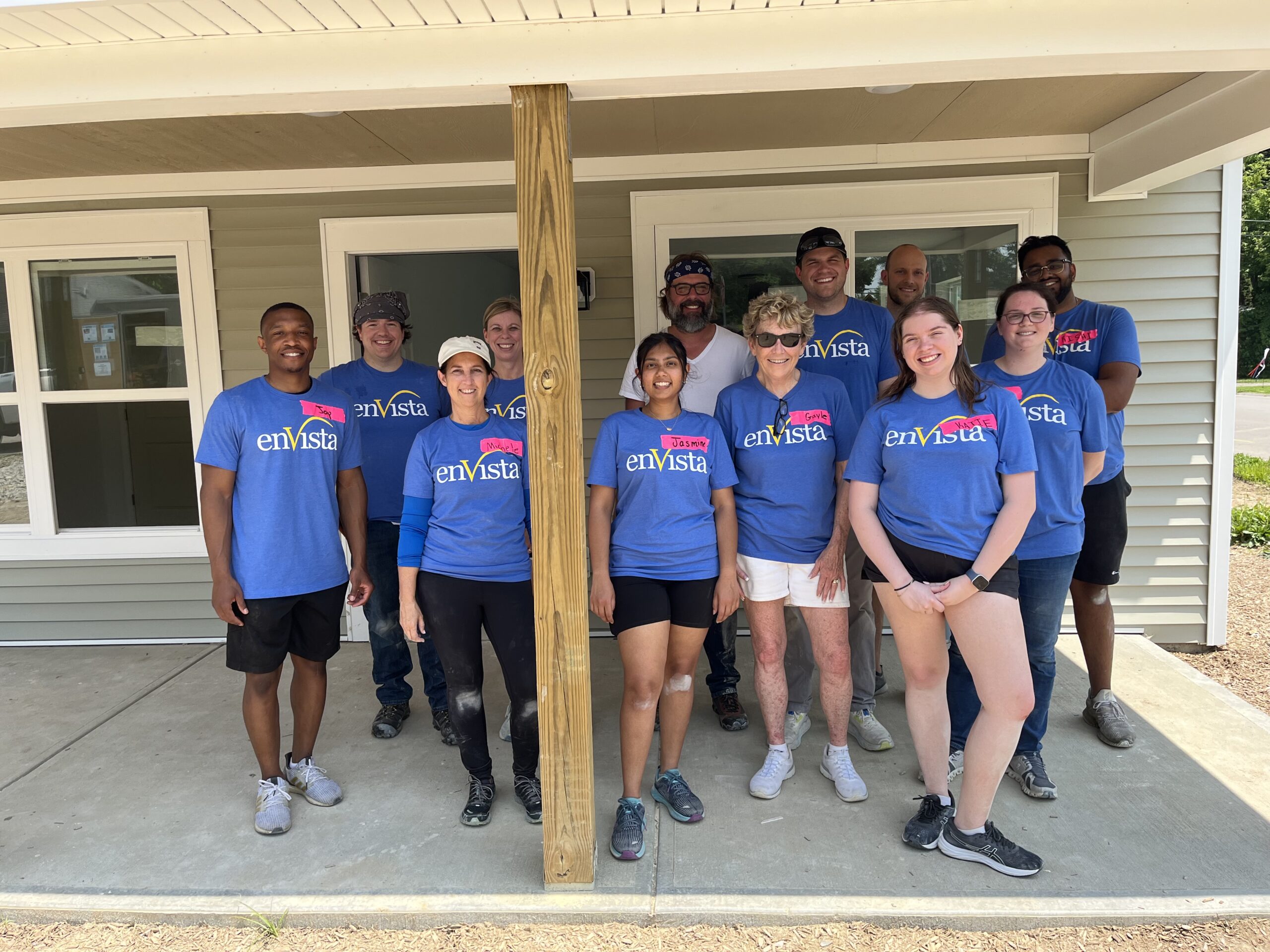A group of twelve people, most wearing matching blue “enVista” T-shirts, stand smiling in front of a house with light siding, posing for a group photo on a sunny day.