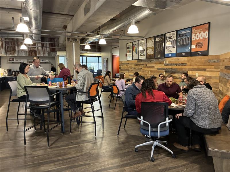 Office workers gather in a break room, eating and socializing at tables. The room has wooden walls, posters, and a counter with food. People chat in groups, creating a casual, lively atmosphere.