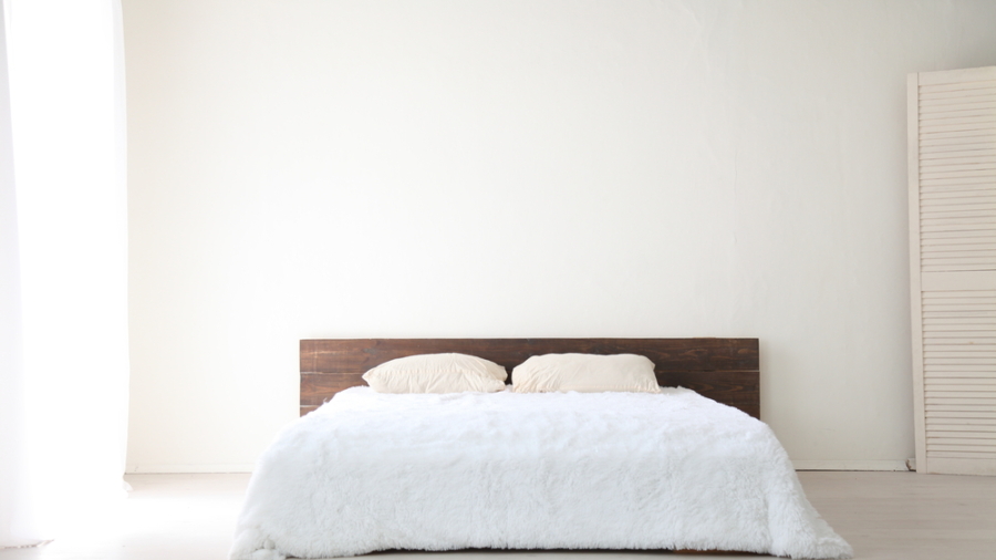 A minimalist bedroom with a low wooden bed frame, part of the Warehouse Advantage Library collection, features two beige pillows and a white fluffy comforter against a plain white wall. Soft natural light streams in, highlighting the cream rug on the floor.