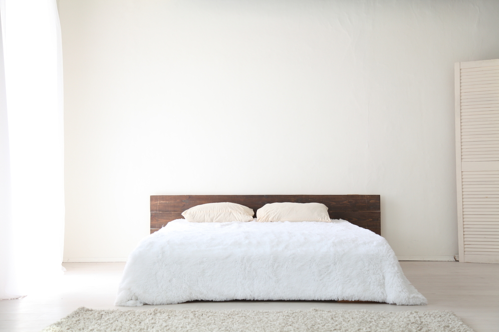 A minimalist bedroom with a low wooden bed frame, part of the Warehouse Advantage Library collection, features two beige pillows and a white fluffy comforter against a plain white wall. Soft natural light streams in, highlighting the cream rug on the floor.