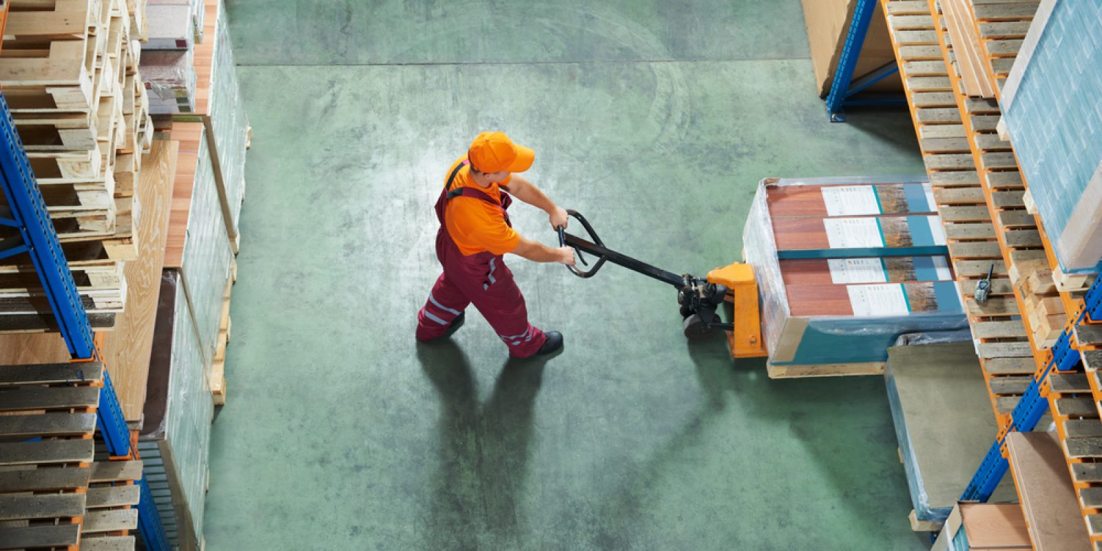 A warehouse worker in an orange shirt and cap uses a pallet jack to move a wrapped pallet between tall 3PL shelving units stocked with goods. The scene is viewed from above.