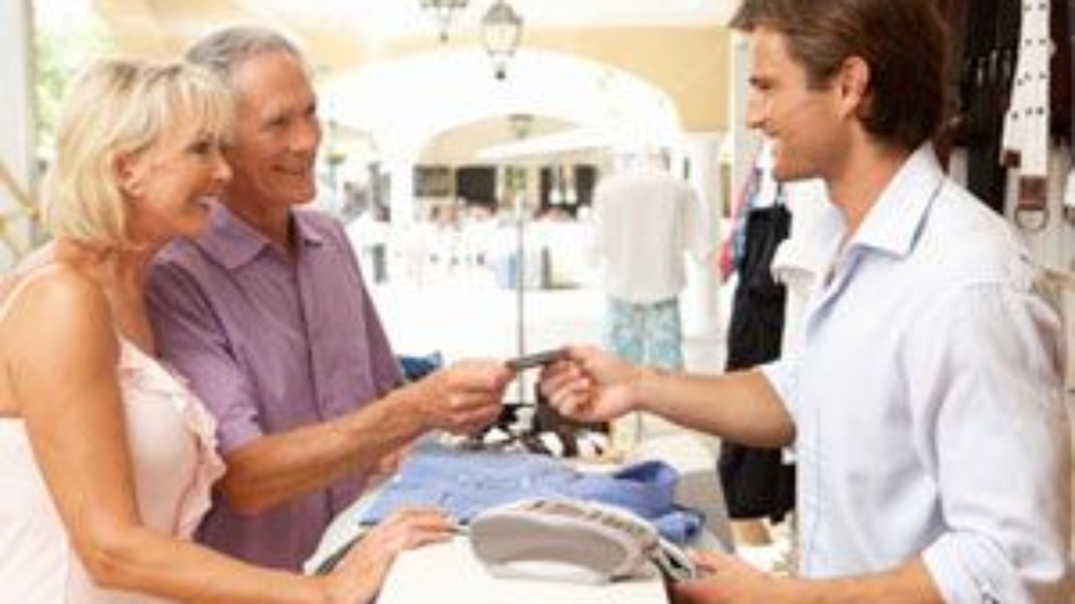 An older couple stands at a store counter, smiling as the man hands a credit card to a young male cashier. Clothing items and a card payment terminal are on the counter. The setting appears bright and welcoming.