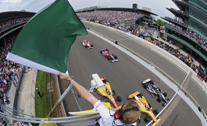 ERP-upgrade A race official waves a green flag to start an IndyCar race as several race cars speed past the starting line on a track, with a large crowd in the grandstands.