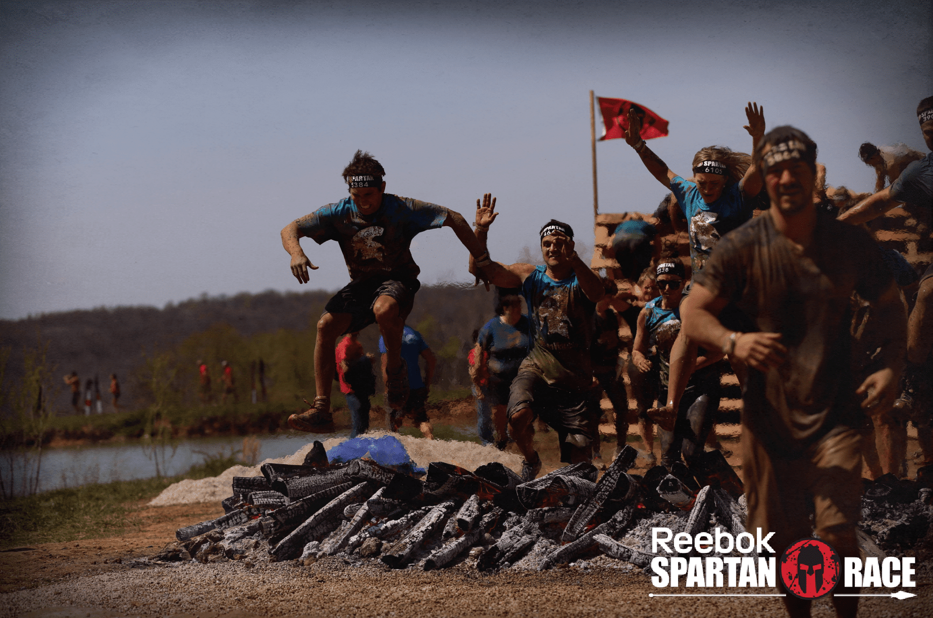 FireZone3 (1) A group of athletes wearing headbands and muddy clothes jump over flaming logs during a Reebok Spartan Race obstacle course outdoors. The event logo is visible in the bottom right corner.