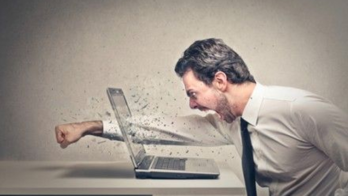 A man in a dress shirt and tie angrily punches a laptop on a desk, causing the screen to shatter and pieces to fly outward.