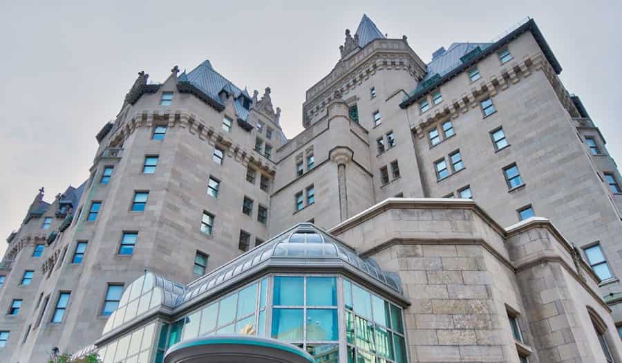 A view looking up at a large, historic stone building with turrets and steep blue-gray roofs, featuring many windows and a modern glass entrance in the foreground.
