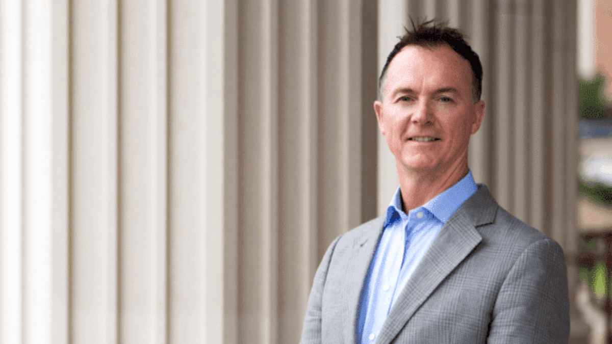 A man in a gray suit jacket and blue shirt stands in front of tall, white columns, smiling at the camera.