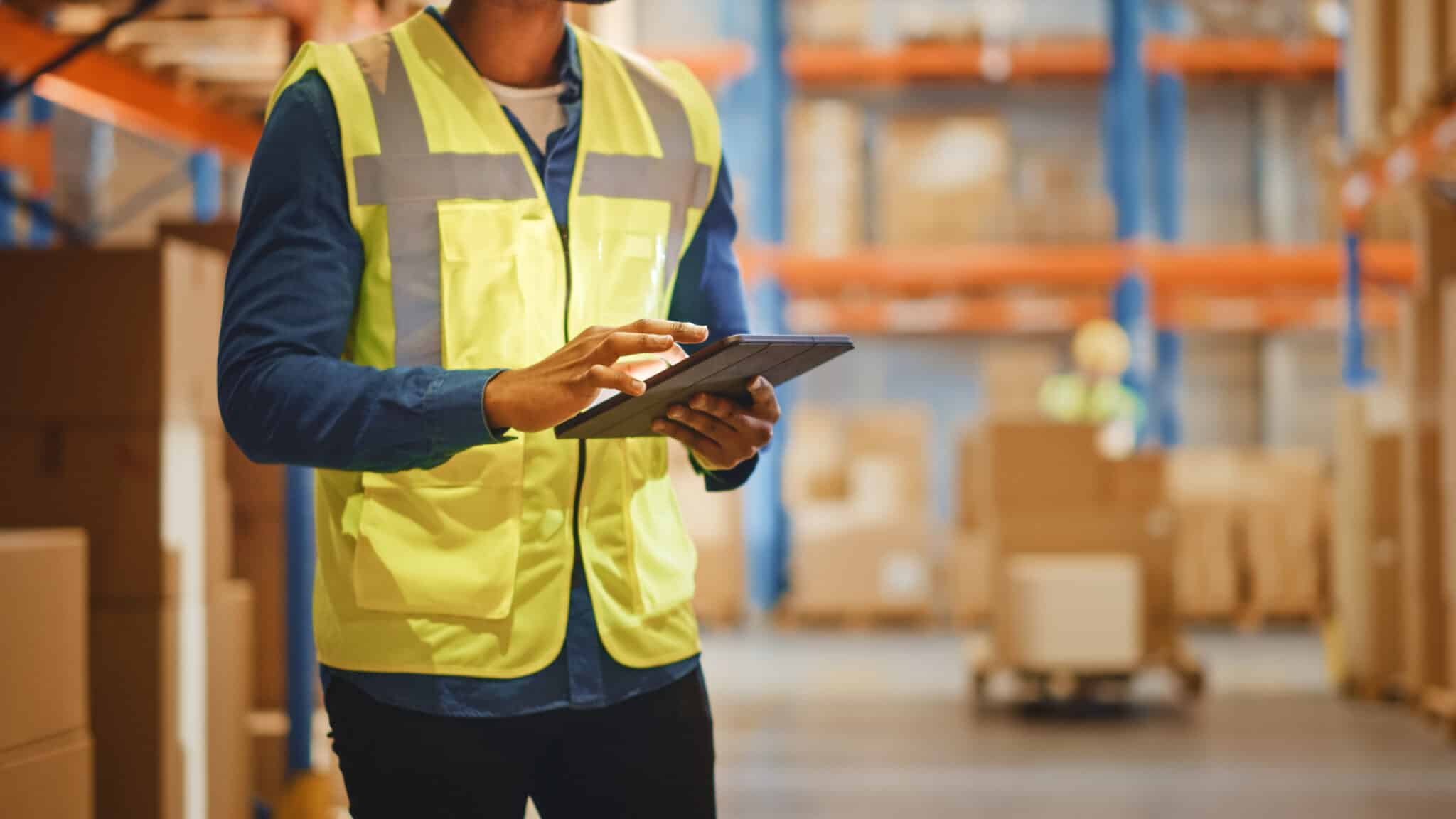 A person wearing a yellow safety vest uses a tablet in a warehouse filled with shelves and cardboard boxes.