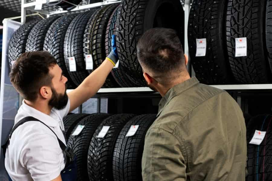 Two men examine and discuss tires displayed on shelves in a store; one man wearing a glove points at a tire while the other listens, with rows of tires and price tags visible.