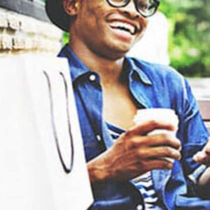 A smiling person wearing glasses and a hat sits on a bench holding a coffee cup and a smartphone, with shopping bags beside them. The person appears relaxed and happy, possibly enjoying a day out.