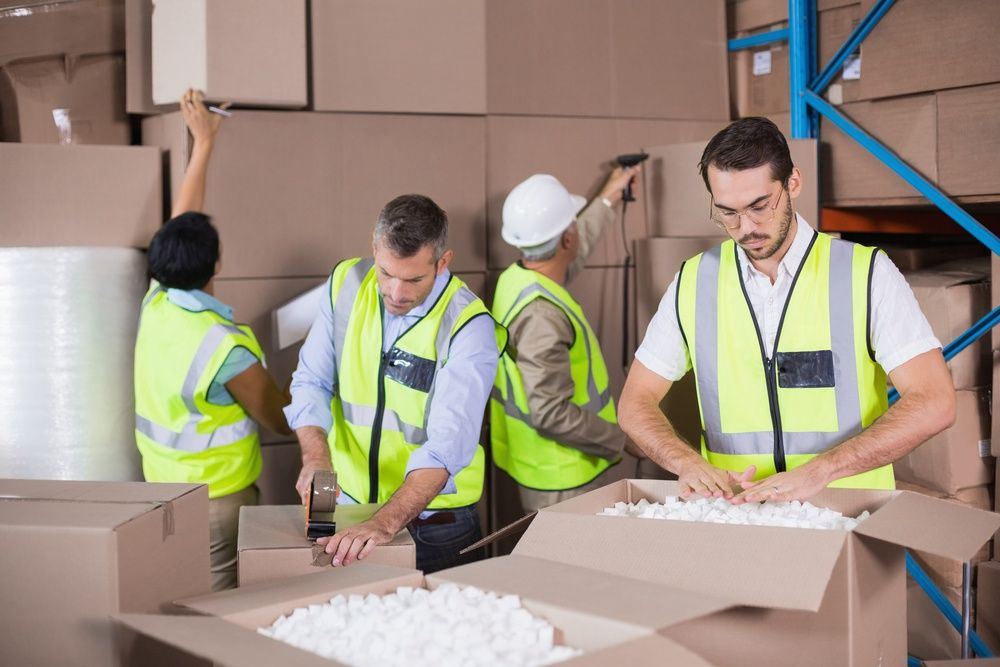 Four workers in high-visibility vests pack and organize cardboard boxes in a warehouse. One fills a box with packing material, another tapes a box, while others arrange boxes on shelves.