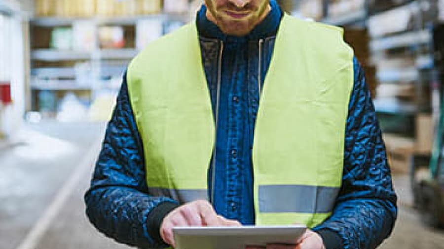 A man wearing a neon yellow safety vest uses a tablet in a warehouse setting, with shelves and boxes blurred in the background.