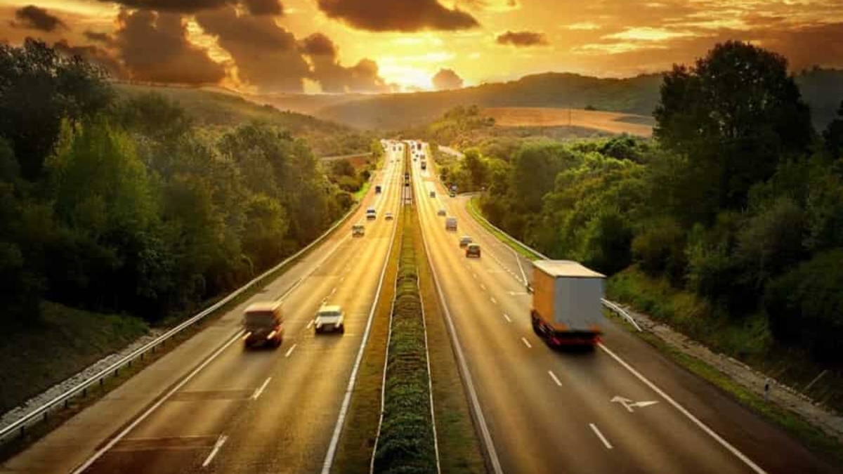 A highway stretches into the distance with cars and trucks driving in both directions, surrounded by green trees and hills, under a dramatic golden sunset sky with clouds.