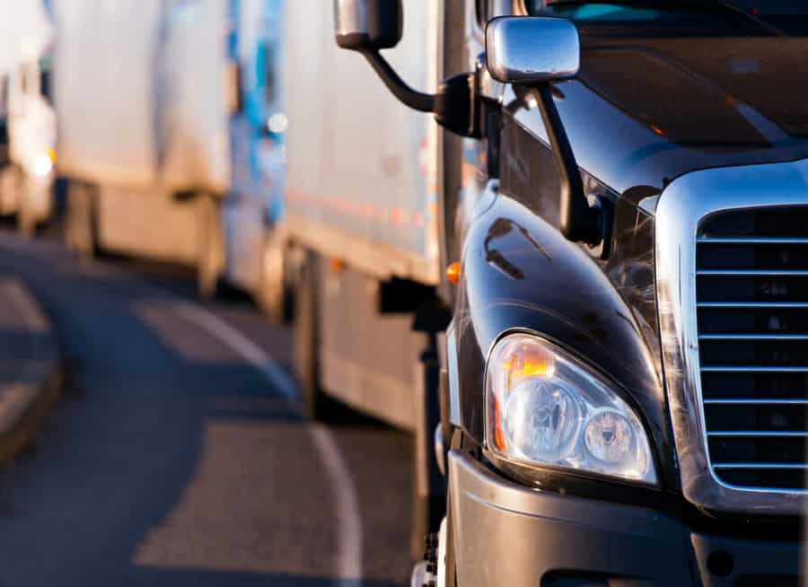 Close-up view of the front of a black semi truck on a curved road, with other trucks in a line ahead, suggesting traffic or a convoy. The scene is bathed in warm sunlight.