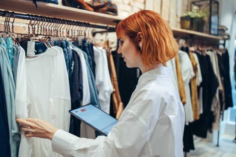 A person with short red hair wearing a white shirt uses a tablet while inspecting clothes hanging on racks in a clothing store.
