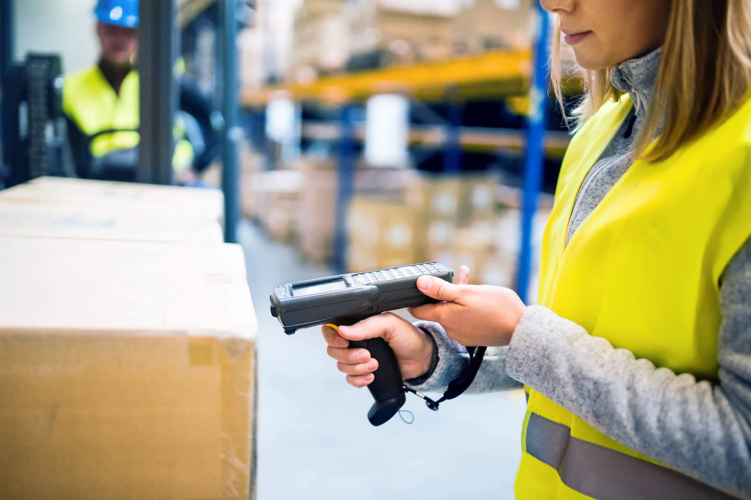 A worker in a yellow safety vest scans a cardboard box with a handheld barcode scanner in a warehouse. Shelves with more boxes and another worker in the background are visible.
