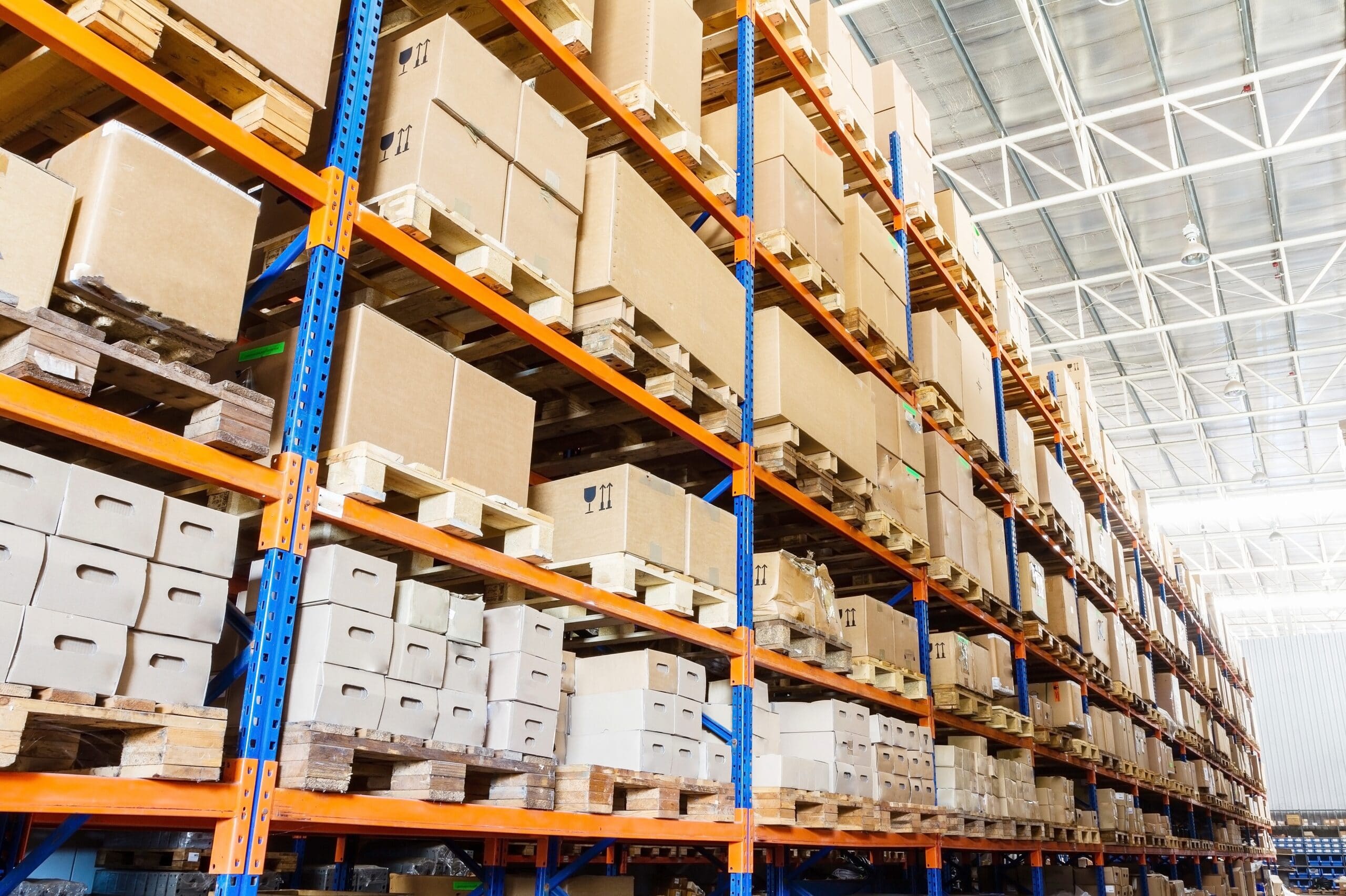 shutterstock_127809008 Rows of tall warehouse shelves filled with stacked cardboard boxes and packages, organized on wooden pallets in a brightly lit industrial storage facility.