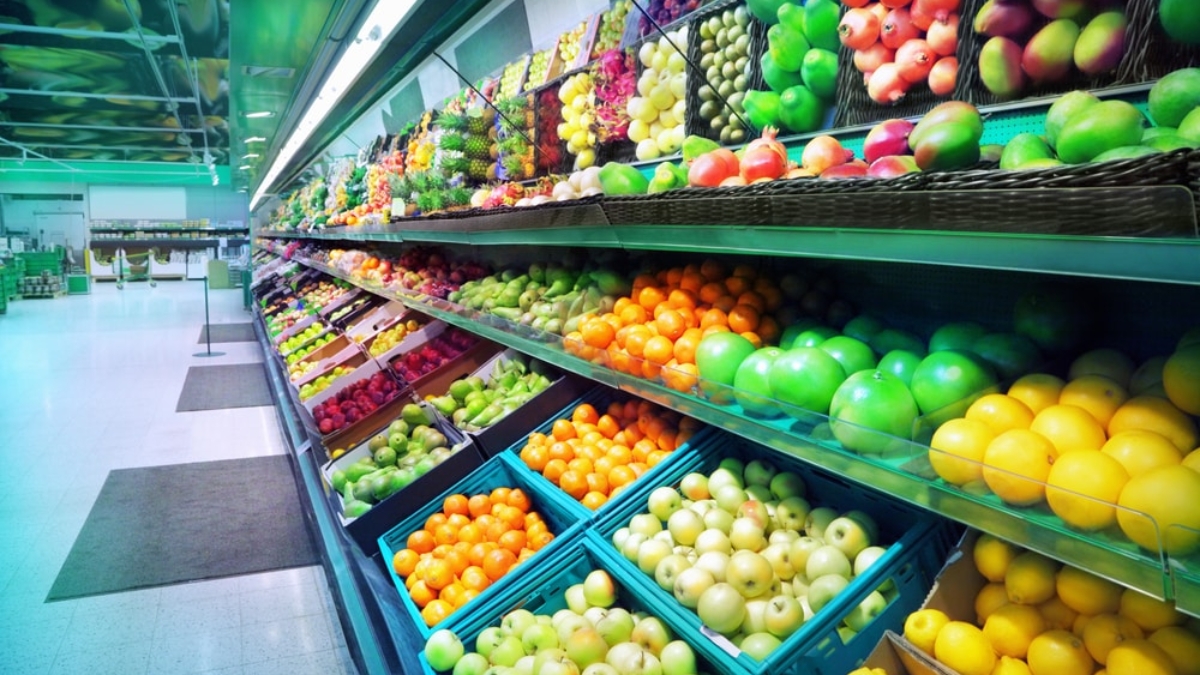 Shelves in a grocery store filled with a colorful variety of fresh fruits and vegetables, including apples, oranges, lemons, and peppers, neatly arranged in rows and baskets.