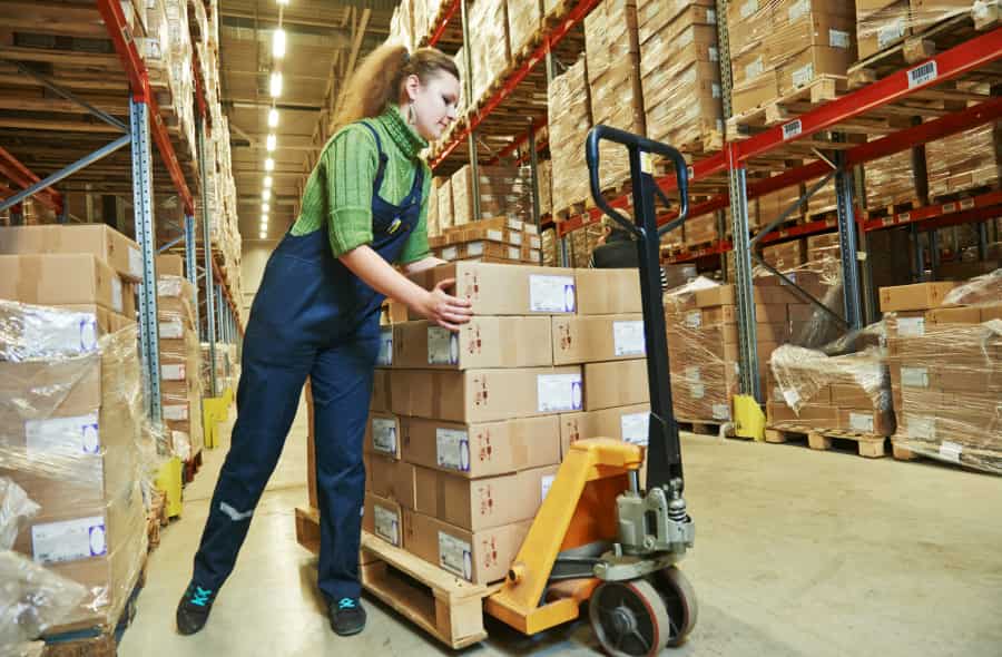A woman in work overalls moves a pallet stacked with cardboard boxes using a pallet jack in a warehouse filled with shelves of boxed goods.