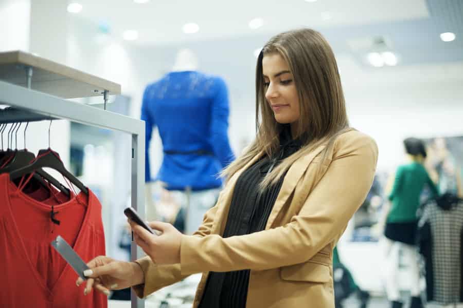 A woman in a tan blazer stands in a clothing store, holding her phone and a price tag while looking at a red dress on a rack. Mannequins and other customers are visible in the background.