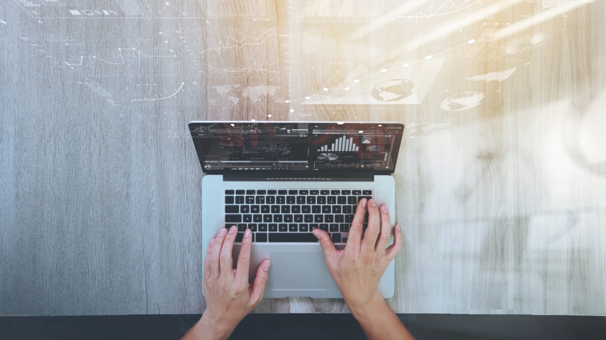 A person typing on a laptop at a wooden desk, viewed from above, with data graphs and charts digitally overlaid on the image, suggesting analysis or business work.