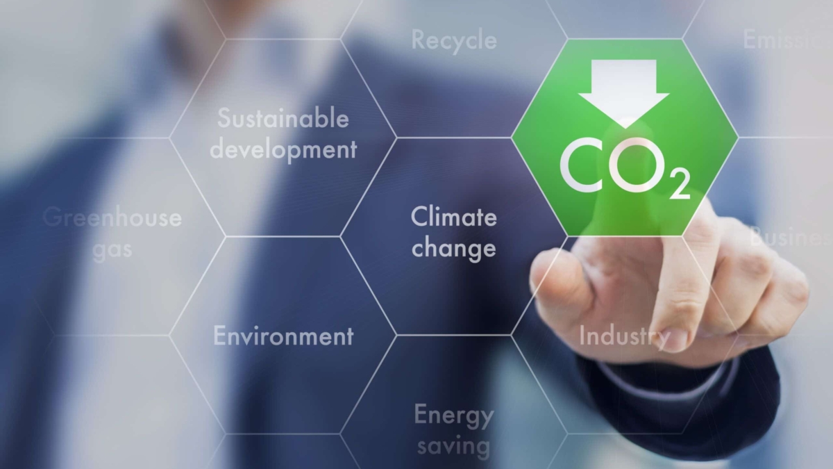 A person presses a digital hexagon labeled CO₂ with a downward arrow on a transparent screen. Surrounding hexagons display words like Climate change, Sustainable development, and Environment.