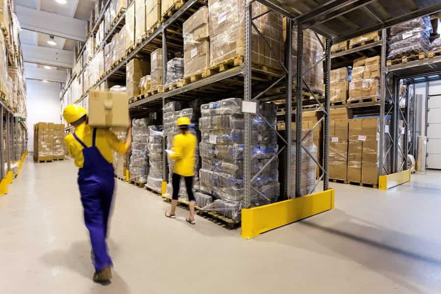 Workers in yellow safety helmets and uniforms move boxes in a large warehouse filled with stacked pallets, shelves, and packaged goods, with high ceilings and bright lighting.