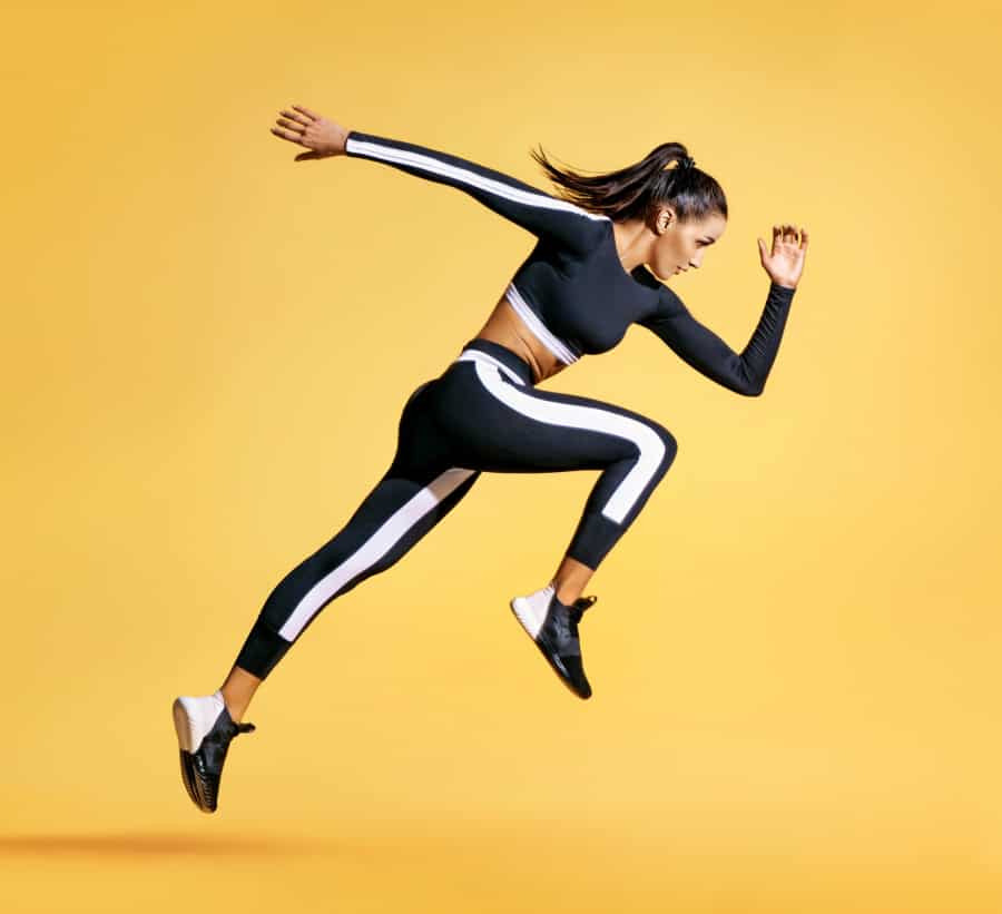 Woman in black and white athletic wear jumps in mid-air against a solid yellow background, appearing energetic and focused, with her arms and legs extended in a running pose.