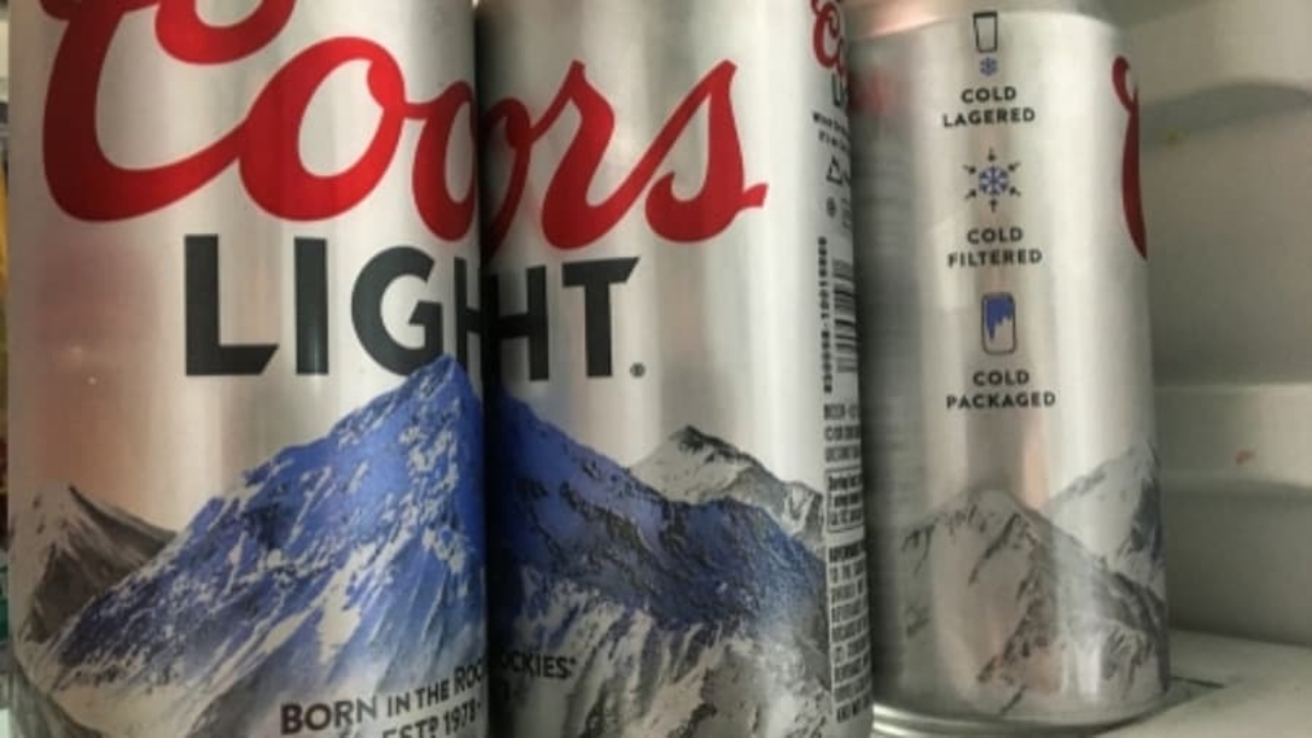 Three Coors Light beer cans with a mountain design are standing in a refrigerator. The cans are silver with red and blue accents, and the Coors Light logo is prominently visible on the front can.