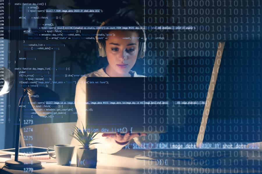 A woman wearing headphones works on a laptop at a desk, overlaid with blue computer code and binary numbers, suggesting programming or cybersecurity activity in a modern workspace.