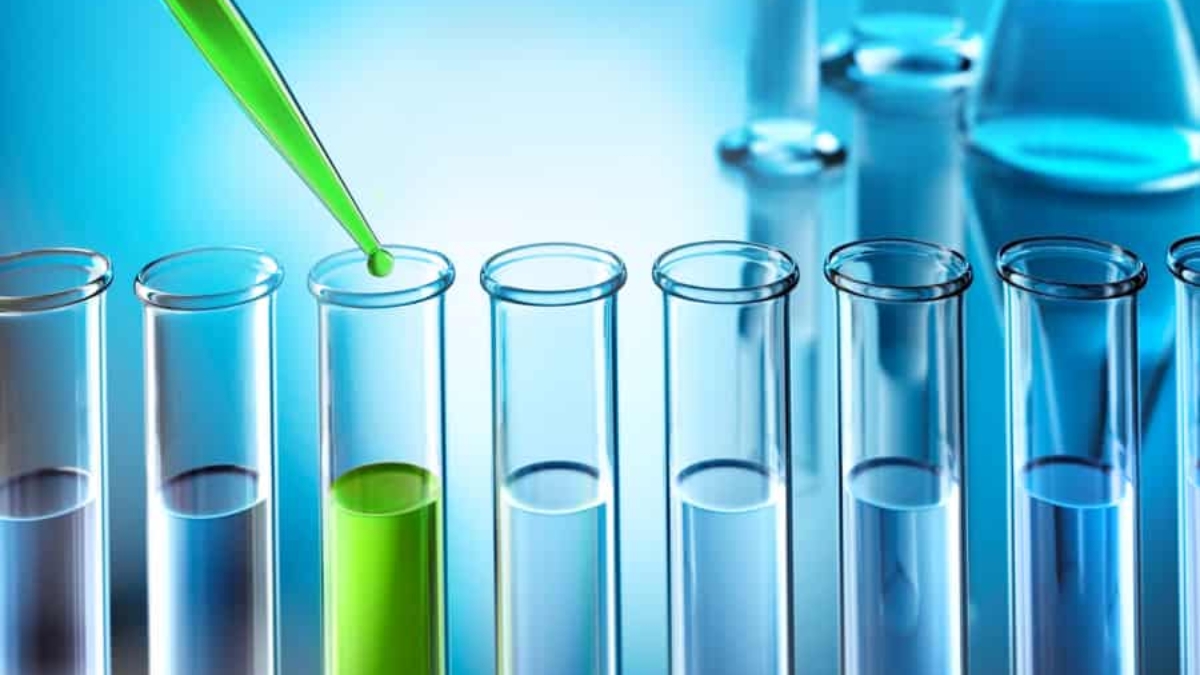 A green liquid is being dropped from a pipette into one of several clear test tubes, with the surrounding lab equipment blurred in the blue-toned background.