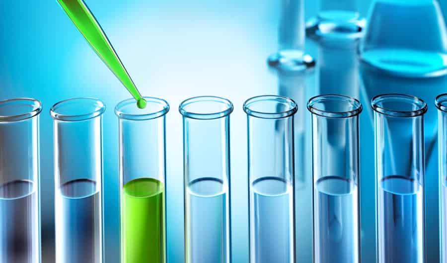 A green liquid is being dropped from a pipette into one of several clear test tubes, with the surrounding lab equipment blurred in the blue-toned background.