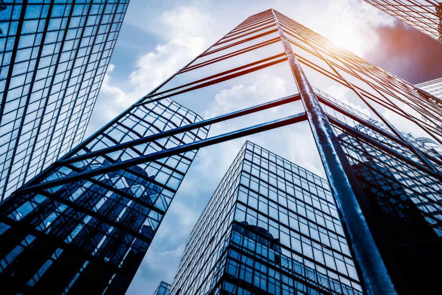 Tall glass skyscrapers reflect the blue sky and clouds as sunlight streams between the buildings, creating an urban cityscape with a modern architectural feel.