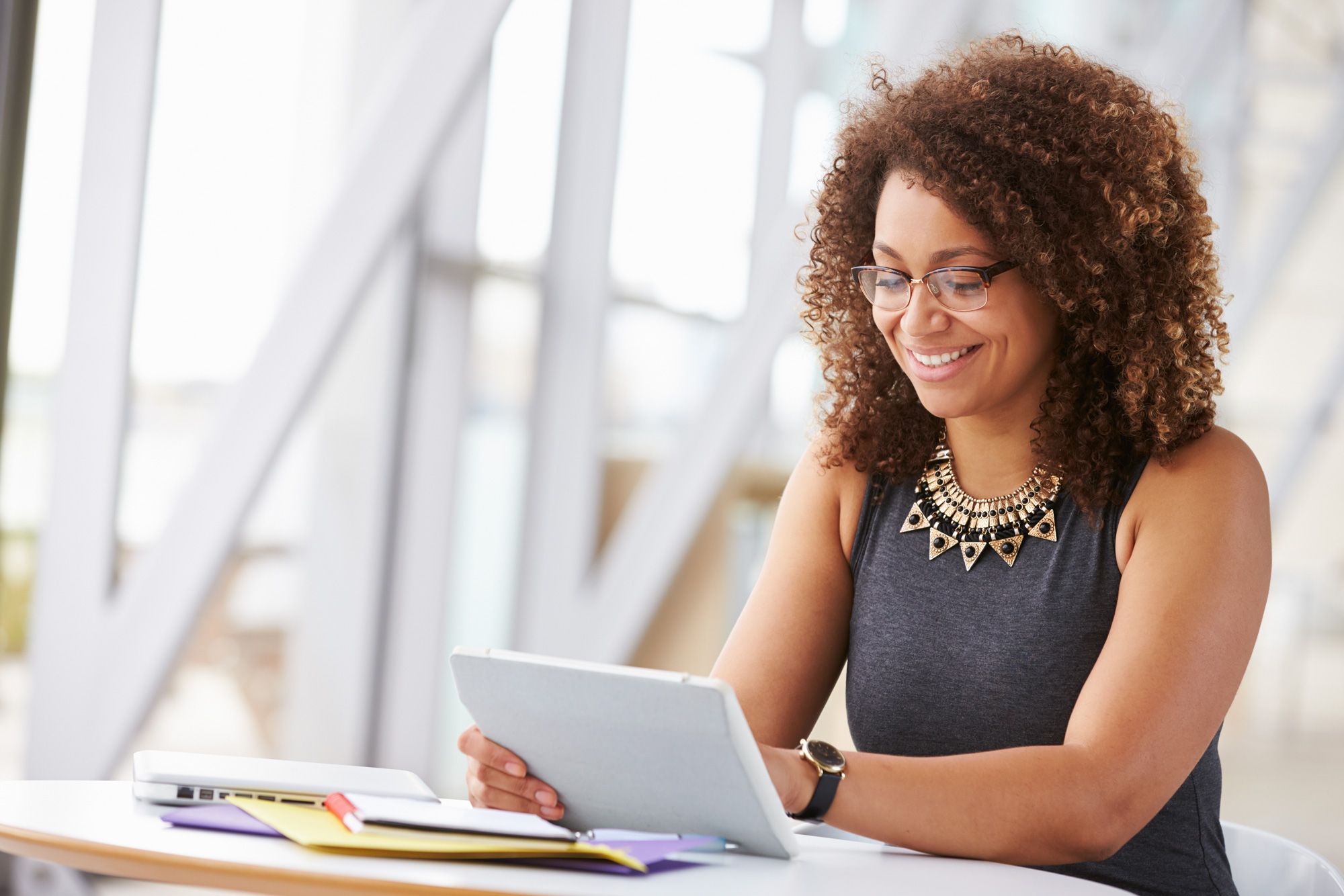power_platform_bg A woman with curly hair and glasses sits at a desk, smiling while looking at a tablet. Papers, a notebook, and a laptop with power platform dashboards open are on the desk in front of her. The background shows a bright, modern interior.